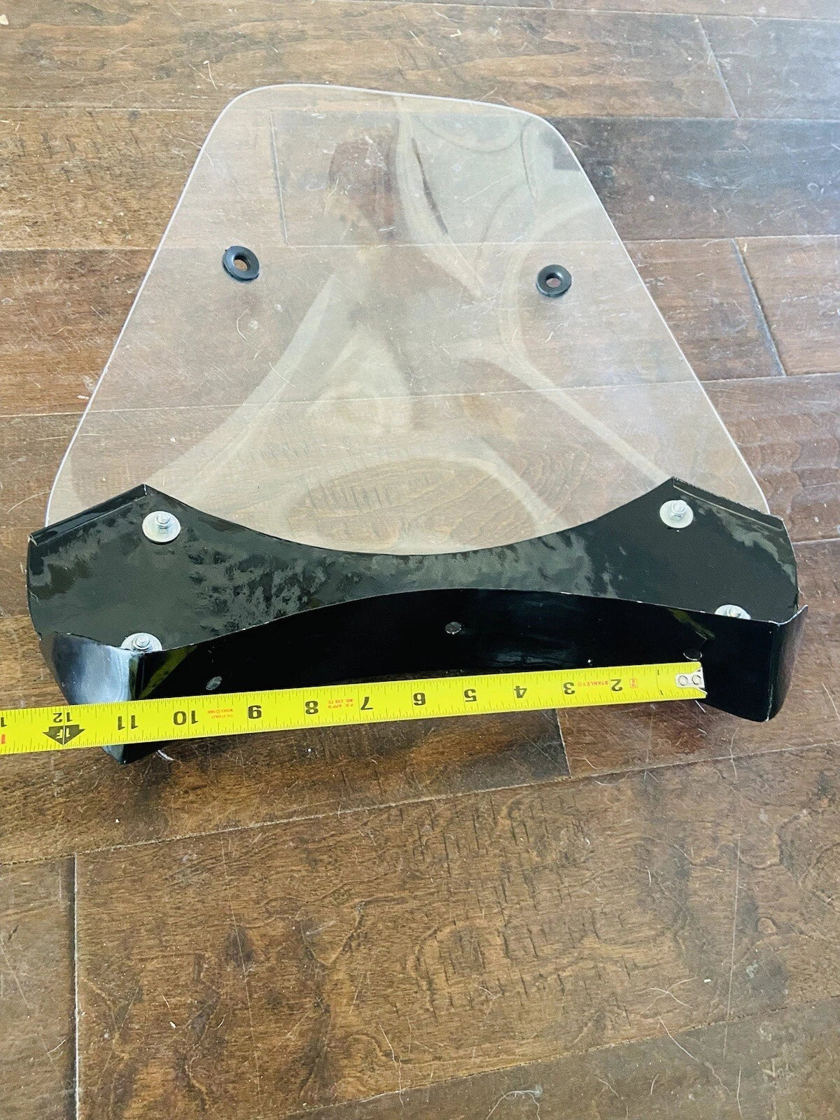 Clear motorcycle windshield with black frame on a wooden floor, measured with a tape measure.