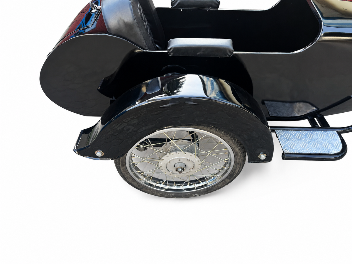 Close-up of a black VOX motorcycle sidecar with chrome spoke wheel, drum hub, black fender, armrest, and diamond-plate footboards